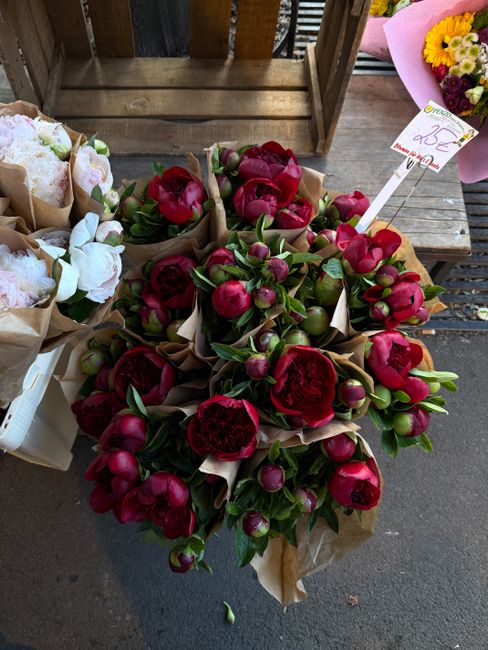 A floral arrangement of large, deep red peonies with lush green foliage, wrapped in brown paper, displayed on a wooden surface at a flower shop. The bouquet appears fresh with full, vibrant petals and