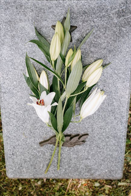 A floral tribute featuring white lilies with large, trumpet-shaped petals and prominent red stamens, along with several closed, elongated buds and long, slender green leaves. The flowers are arranged