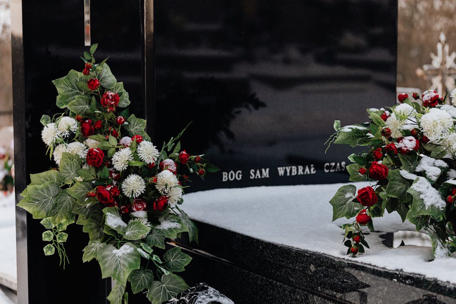 A memorial gravestone adorned with floral arrangements consisting of red roses, white chrysanthemums, and green foliage, including ivy and holly leaves, arranged in two bouquets placed on either side