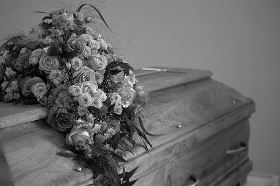 A large funeral floral arrangement resting on top of a polished wooden coffin. The bouquet features a mix of roses and other small flowers in various stages of bloom, with some roses fully open and ot