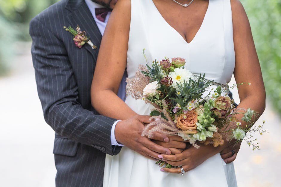 A bride and groom holding a bouquet arrangement of mixed flowers, including soft peach roses, white daisies, green foliage, and delicate pink blooms, wrapped in pale pink tissue paper with a beige rib